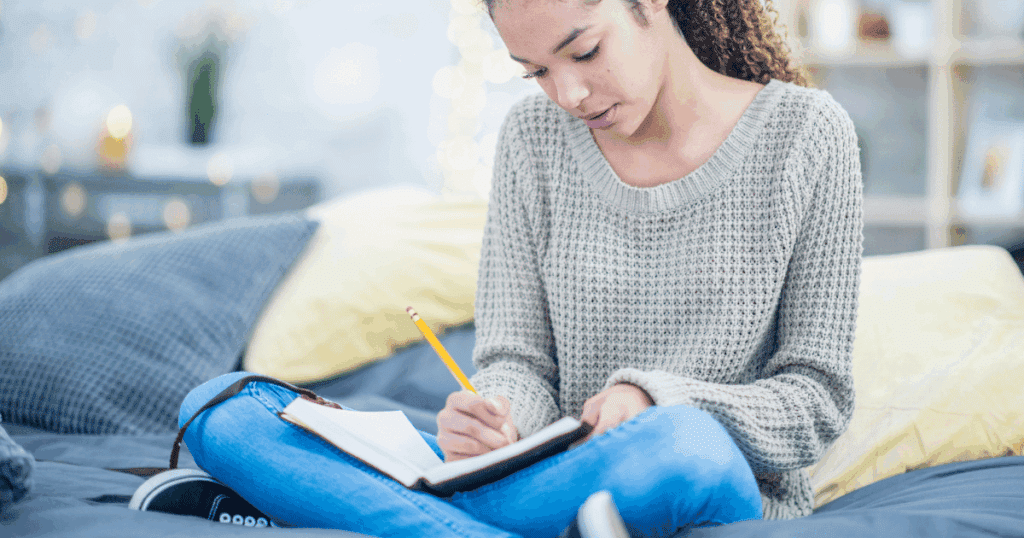 Young woman sitting cross-legged while journaling, focused and peaceful, with a notebook in her lap and a pen in hand, surrounded by a calm, cozy setting.