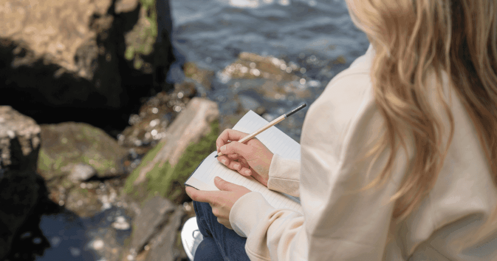 Person sitting by the water, peacefully journaling in a notebook, with a serene view of the lake or ocean in the background, capturing a quiet moment of reflection.